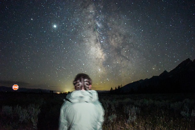 Une jjeune fille de dos regarde le ciel étoilé. Au loin, un panneau stop brille dans la nuit - Photo by Jack Charles on Unsplash