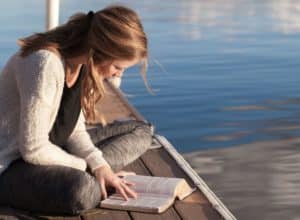 jeune femme lisant la Bible sur un ponton (recadrée) - Bethany Laird sur https://unsplash.com/fr/photos/photo-dune-femme-lisant-un-livre-pres-dun-plan-deau-vGReyBvIX-o