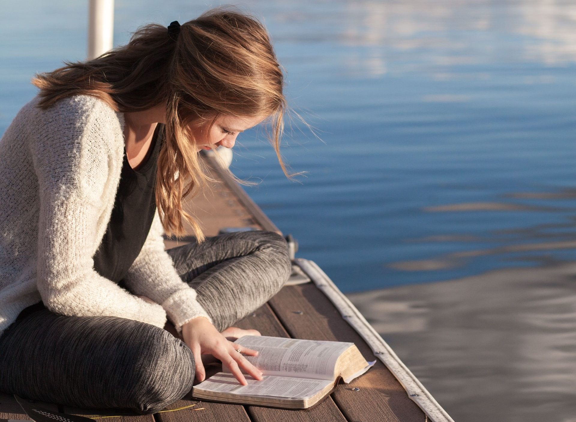jeune femme lisant la Bible sur un ponton (recadrée) - Bethany Laird sur https://unsplash.com/fr/photos/photo-dune-femme-lisant-un-livre-pres-dun-plan-deau-vGReyBvIX-o