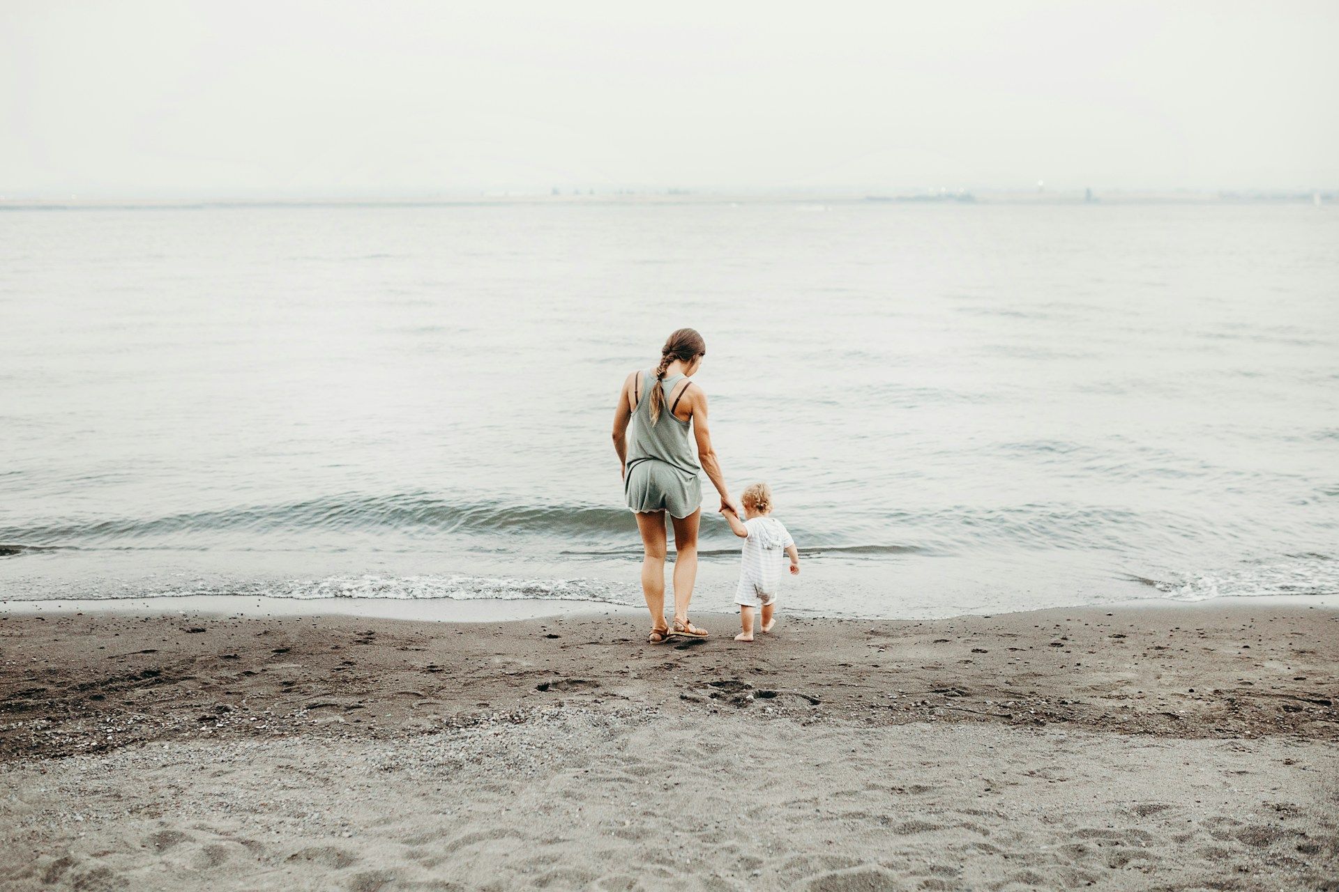 Une mère au bord de la mer tenant par la main un tout jeune enfant - Photo by Liana Mikah on https://unsplash.com/fr/photos/femme-et-bebe-marchant-sur-le-rivage-de-sable-gris-pendant-la-journee-J5DIOISkDko