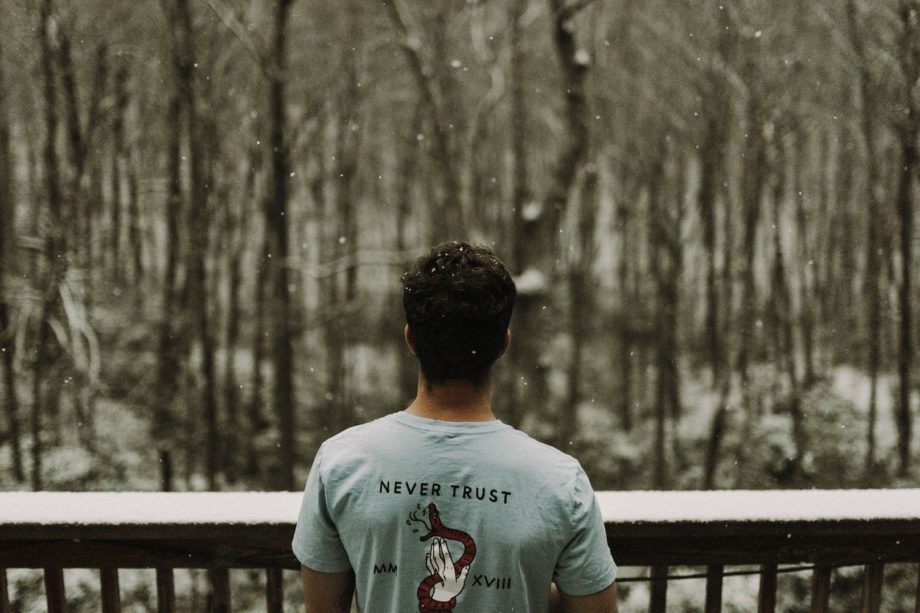 Un homme regarde la neige tomber, avec un tee-shirt évoquant le serpent de la tentation - Photo by Scott Broome on https://unsplash.com/fr/photos/homme-portant-un-t-shirt-gris-debout-a-cote-dune-balustrade-en-bois-marron-pendant-la-journee-enneigee-jxvigoarqrQ