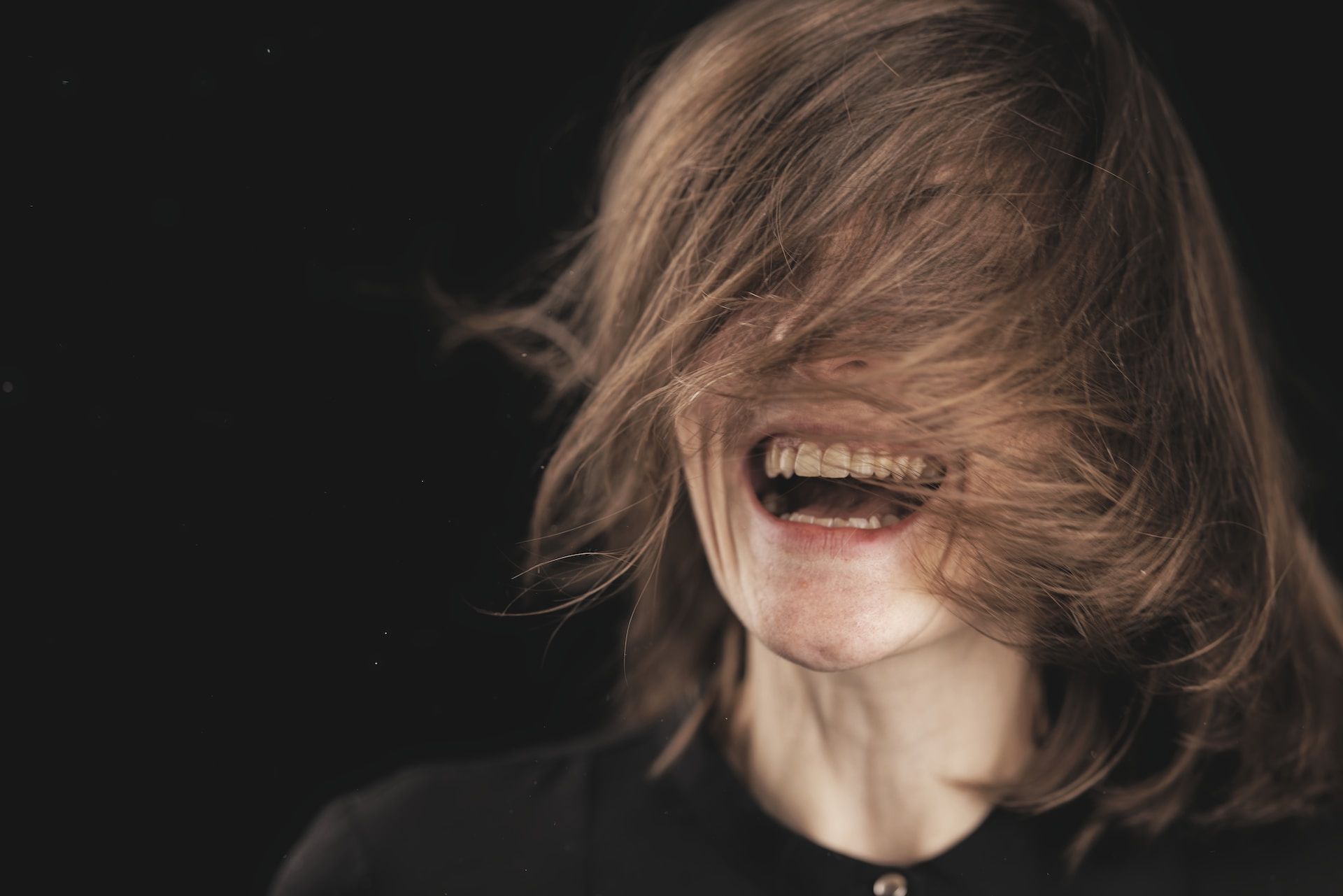 jeune femme riant à pleine dents, le visage caché par ses cheveux - Photo by engin akyurt on https://unsplash.com/fr/photos/femme-en-chemise-noire-a-col-rond-7XzgDjkspmQ