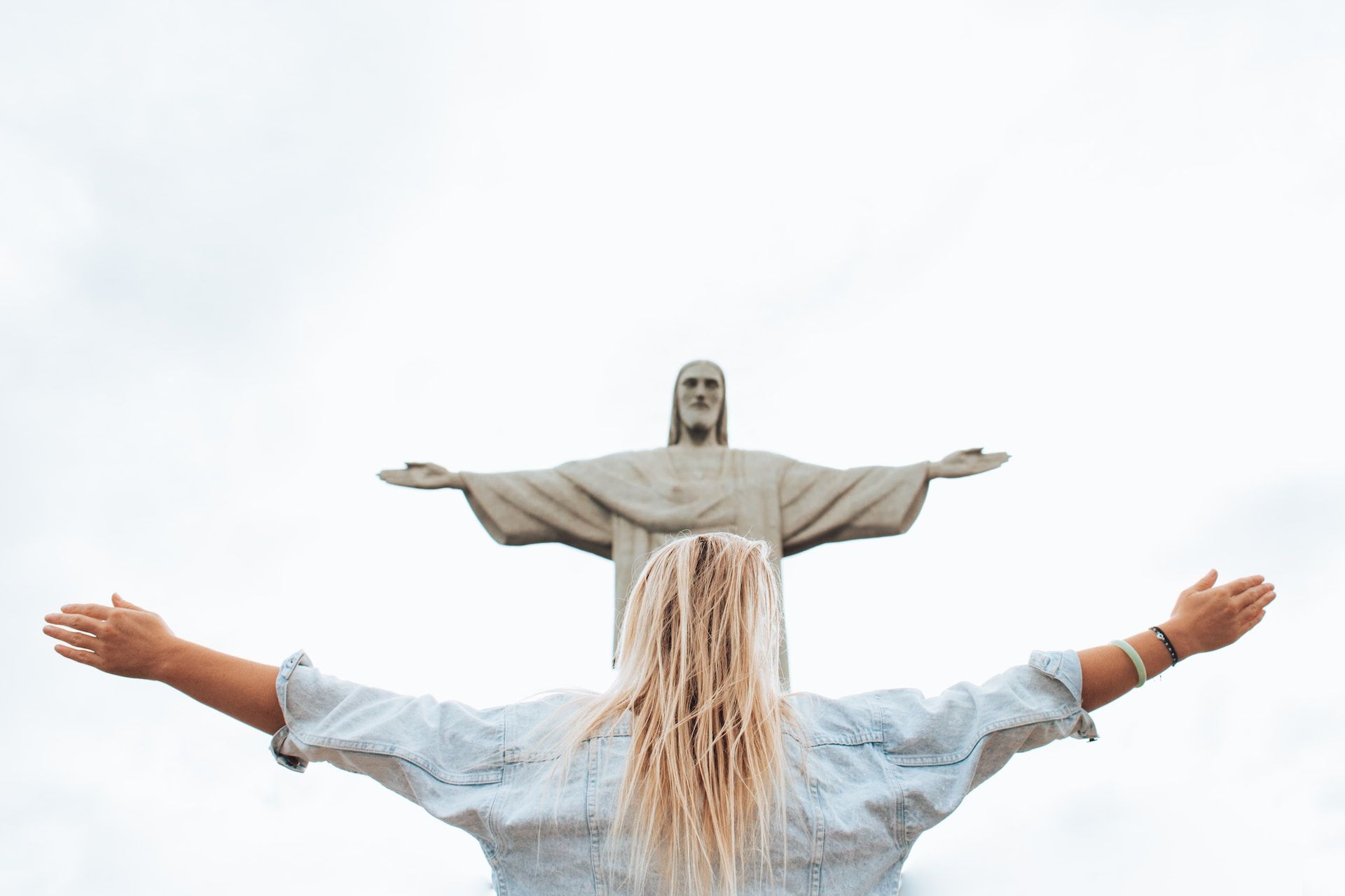 une personne de sexe indéterminé, avec des cheveux longs prend la posture du Christ de Rio - Photo by Marcos Paulo Prado on https://unsplash.com/fr/photos/femme-en-chemise-a-manches-longues-blanche-et-jean-en-denim-bleu-Iv0mvJx9uLM
