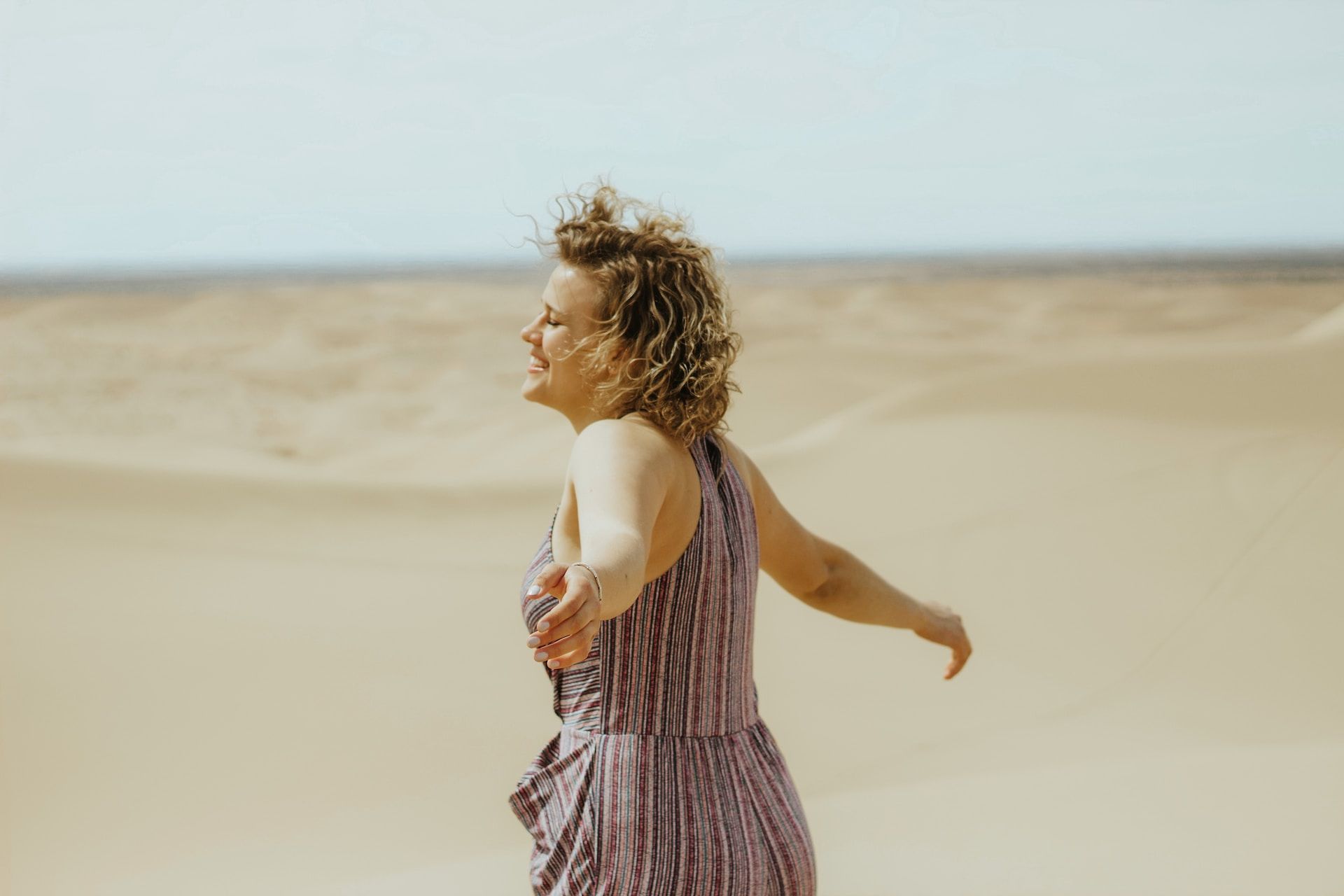 femme sur la place écartant les bras et prenant l'air marin sur son visage - Photo by Tori Wise on https://unsplash.com/fr/photos/femme-en-robe-debardeur-a-rayures-blanches-et-violettes-debout-sur-le-sable-brun-pendant-la-journee-h66XccaKuyI