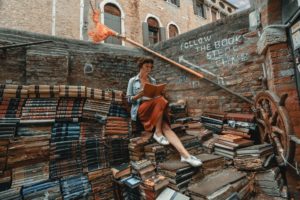 femme lisant, assise sur une pile de livres, avec une inscription derrière. Photo by Clay Banks on https://unsplash.com/photos/NGupON6JOYE 