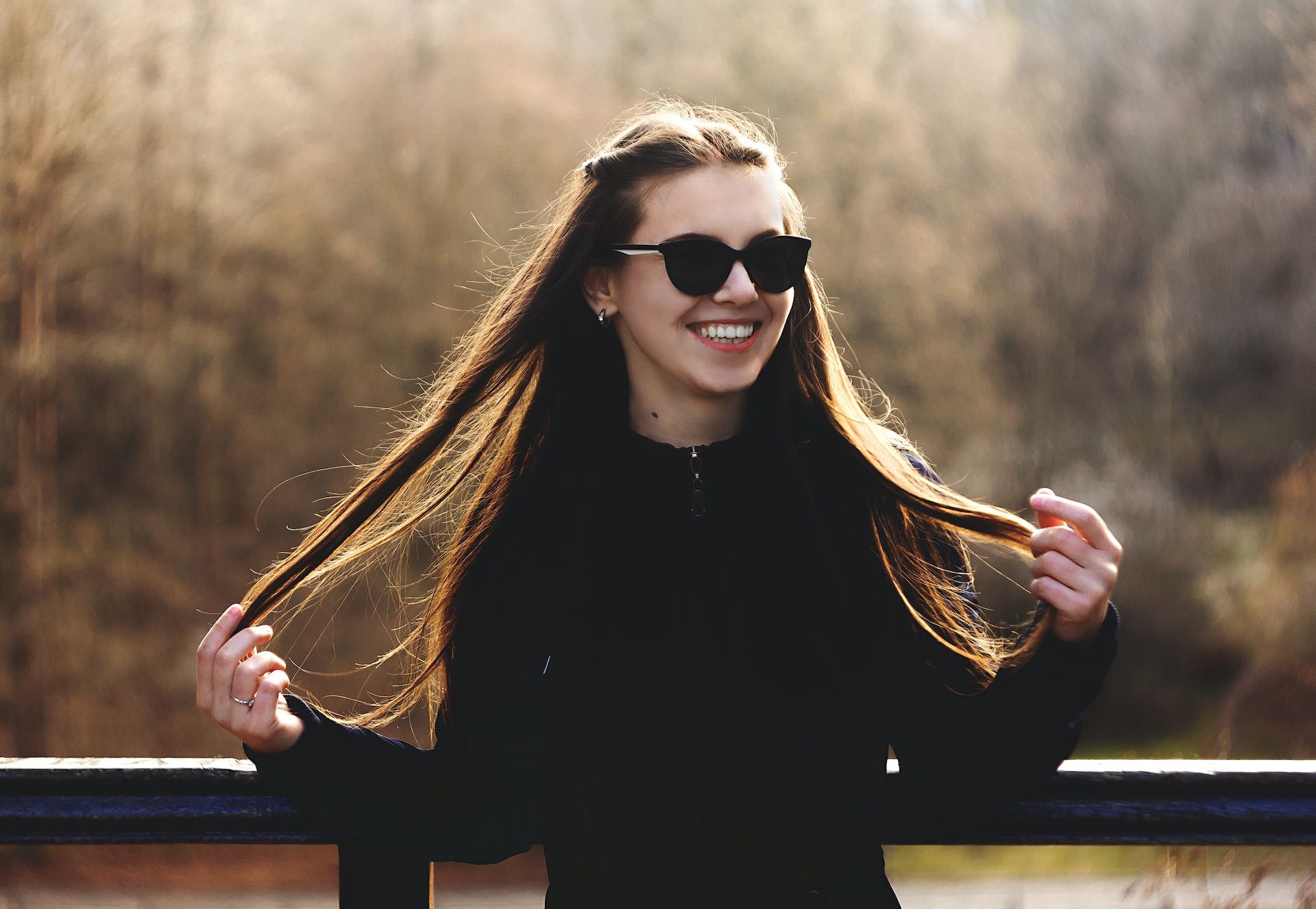 Jeune femme souriante avec des lunettes de soleil sur un balcon, tennat ses cheveux - Photo de Abbat sur https://unsplash.com/fr/photos/WXlEZq8iSyc