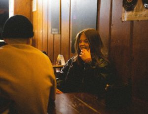 Un couple dans un bar, discutent. La femme est de face et souriante, l'homme est de dos - Photo de BĀBI sur https://unsplash.com/fr/photos/une-femme-assise-a-une-table-parlant-au-telephone-portable-s5mbvU0yias