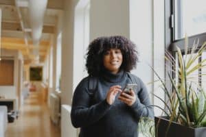 une jeune femme souriante avec son téléphone dans une bibliothèque - Photo de Good Faces sur https://unsplash.com/fr/photos/femme-en-chemise-noire-a-manches-longues-utilisant-un-smartphone-DavJROC1lBM