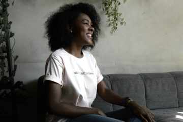 Une femme noire moderne en tee-shirt souriante - Photo de DISRUPTIVO sur https://unsplash.com/fr/photos/femme-souriante-assise-sur-le-canape-DokE5D4GbDk