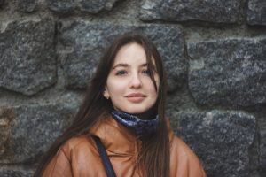 Une jeune femme debout devant un mur de pierre, souriante, nous regarde - Image par Abbat de https://pixabay.com/fr/photos/fille-portrait-maquette-brunette-6093779/