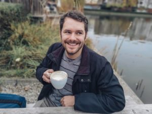 Un homme souriant avec une tasse de café à la main, assis à une table à l'extérieur, près d'un lac - Image par melancholiaphotography de https://pixabay.com/fr/photos/homme-caf%C3%A9-en-plein-air-lac-3803551/