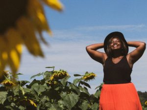 une femme africaine souriante sur un fond de nature - Photo de Eye for Ebony sur https://unsplash.com/fr/photos/femme-en-debardeur-noir-dans-le-champ-de-tournesol-pendant-la-journee-IiTtcSj8lQ8