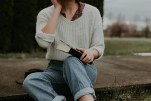 Une jeune femme dont on ne voit pas le visage, en pull blanc et jean, portant une bible fermée - Photo de Priscilla Du Preez 🇨🇦 sur https://unsplash.com/fr/photos/femme-assise-tout-en-tenant-un-livre-jYUvMwABmGs