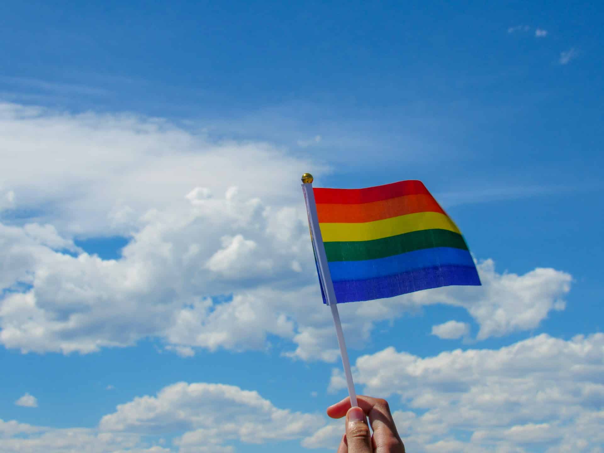 drapeau arc-en-ciel brandi en l'air, sur fond de ciel bleu avec d'agréables nuages blancs Photo de Brielle French sur https://unsplash.com/fr/photos/personne-tenant-le-drapeau-de-lamerique-sous-les-nuages-blancs-et-le-ciel-bleu-pendant-la-journee-61JpASTemRU