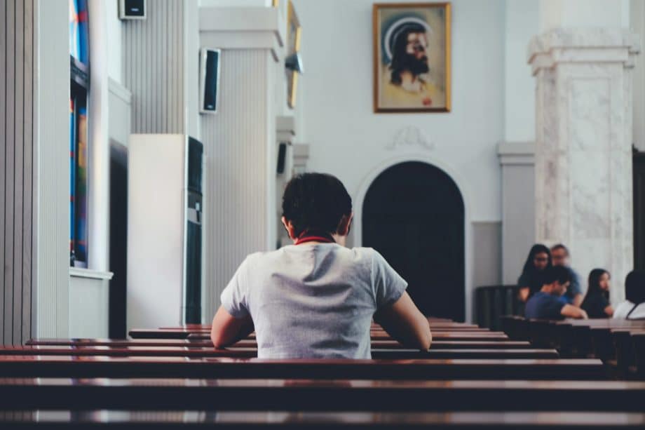 Un jeune dans une église assis sur un banc, avec des personnes plu sloin en arrière plan - Photo de Rimon Mori sur Unsplash