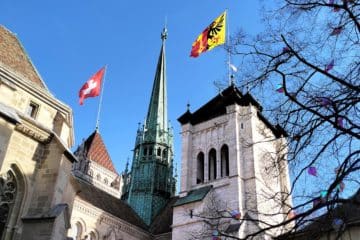 La cathédrale Saint-Pierre, avec les drapeaux de la Suisse et du canton de Genève pavoisant les tours - Photo de Roshand Bhantooa sur https://unsplash.com/fr/photos/une-eglise-avec-un-clocher-et-une-tour-dhorloge-IJJSz4Wvlhw