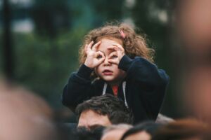 Une petite fille assise sur les épaules de son père regarde au loin en faisant des lunettes avec ses mains - Photo de Edi Libedinsky sur https://unsplash.com/fr/photos/fille-faisant-un-geste-de-la-main-sur-son-visage-1bhp9zBPHVE