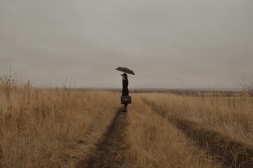 Une femme seule dans un champ par temps gris, portant un parapluie et un cartable de cadre d