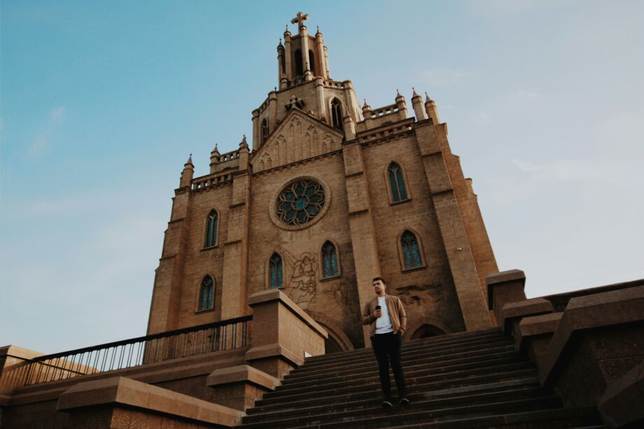Un homme debout sur des marches, surplombé par une église imposante - Photo de Artem Bryzgalov sur https://unsplash.com/fr/photos/homme-debout-sur-les-escaliers-devant-leglise-pendant-la-journee-N8jgY_HDzck